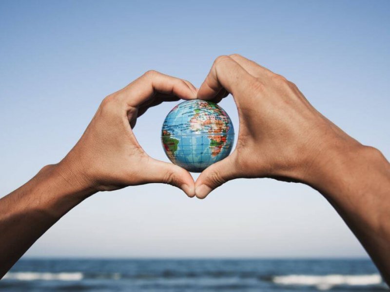 closeup of a young caucasian man with a world globe in his hands forming a heart against the blue sky, in front of the sea