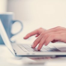 Close up of a womans hands typing on a laptop computer. There is a coffee cup in the background