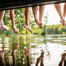 A group of friends legs dangling off a jetty, by the water at a lake together on a sunny afternoon.